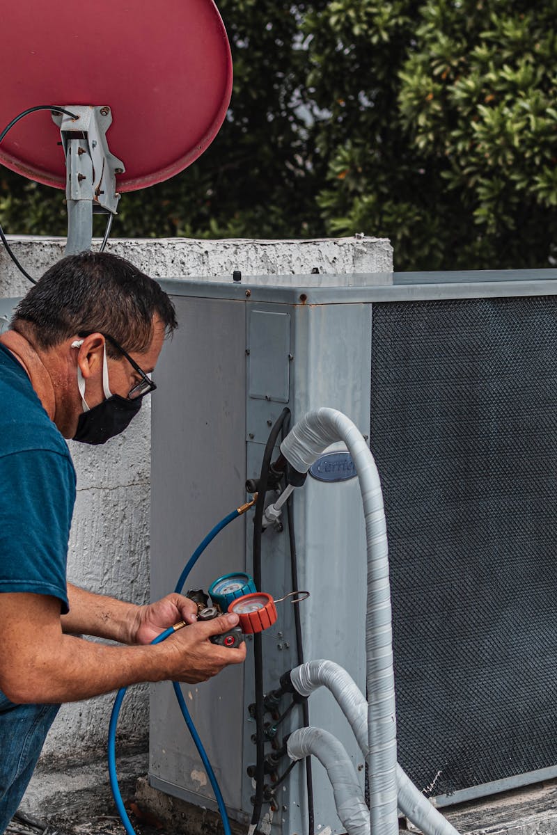 Technician repairing an air conditioner unit outdoors, wearing a facemask and using a manifold gauge.